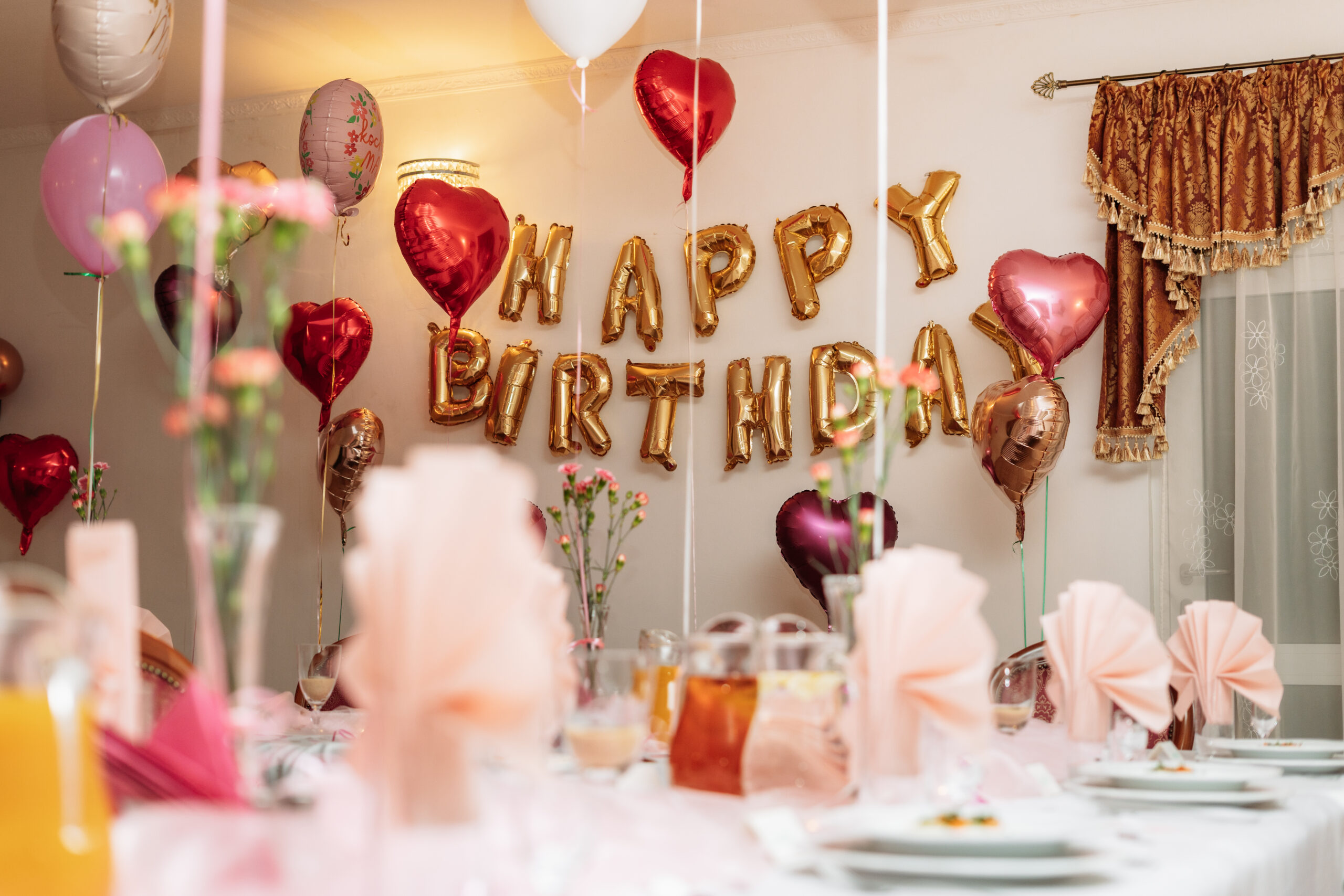 Napis Happy Birthday ze złotych balonów na ścianie, otoczony helowymi balonami w kształcie serc.