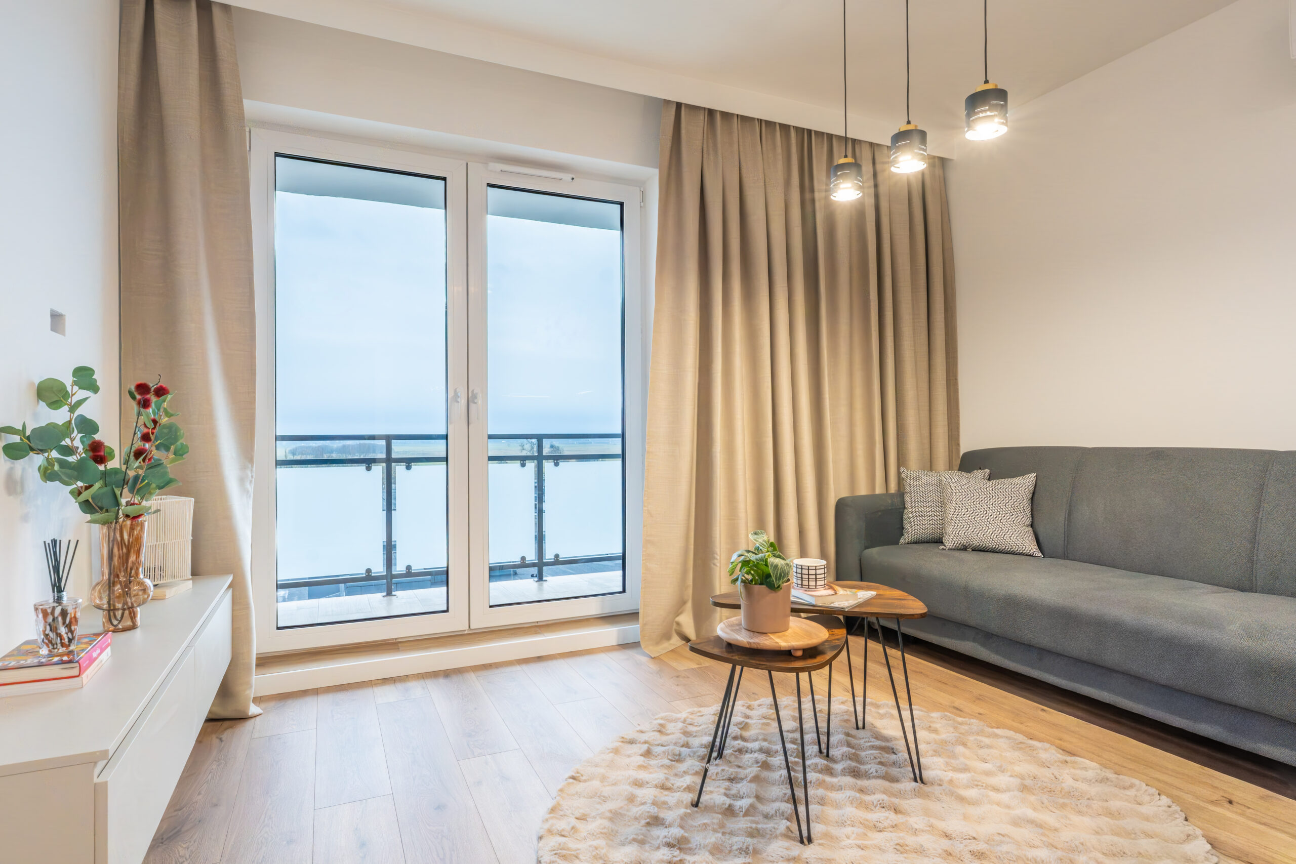 Przestronny, jasny salon z dużą szarą kanapą umieszczoną przy szerokim oknie balkonowym. Przez okno wpada naturalne światło, oświetlając minimalistyczne wnętrze w stonowanej kolorystyce.