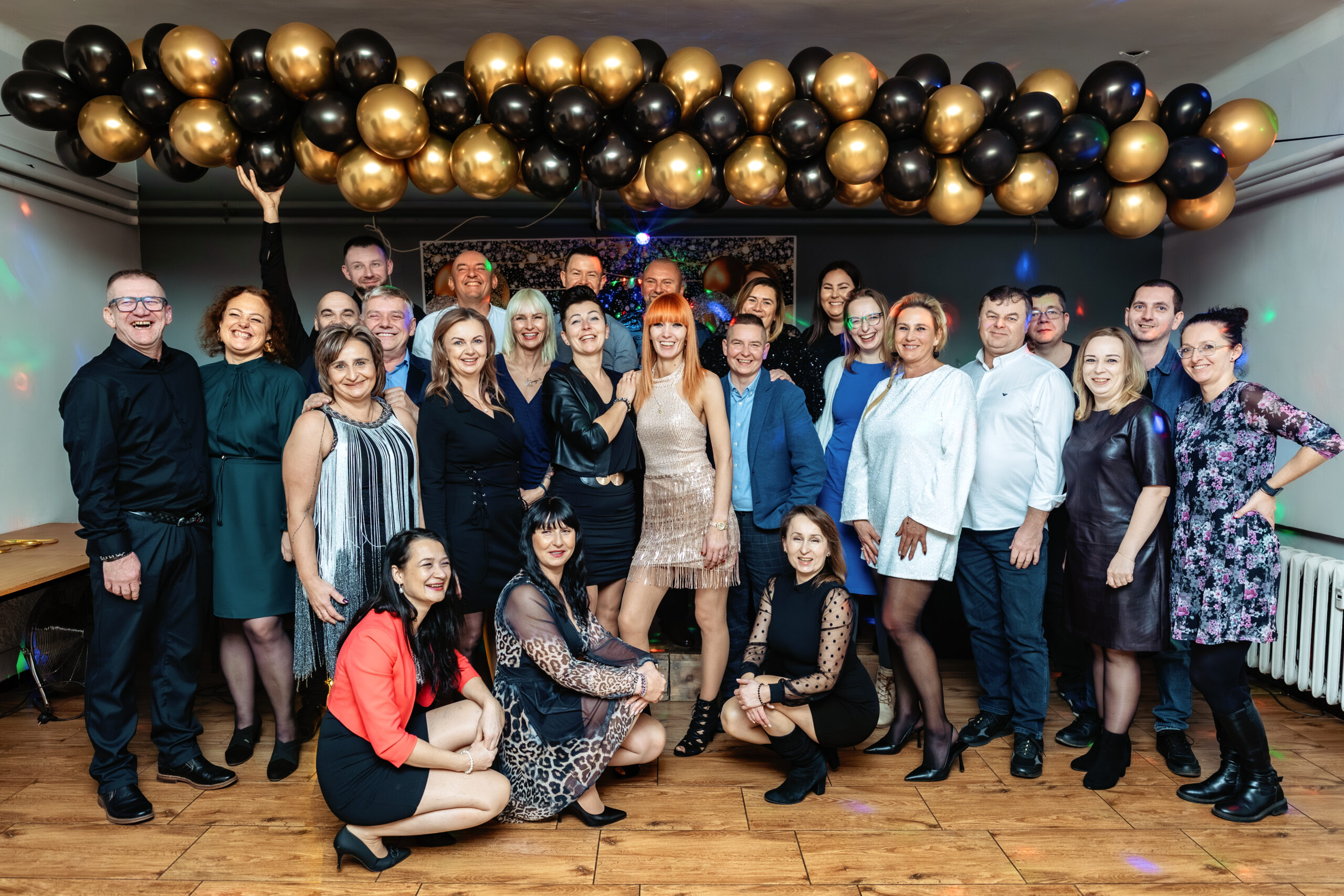 Wielkie zdjęcie grupowe wszystkich gości z jubilatką w centrum, pod girlandą ze złoto-czarnych balonów.