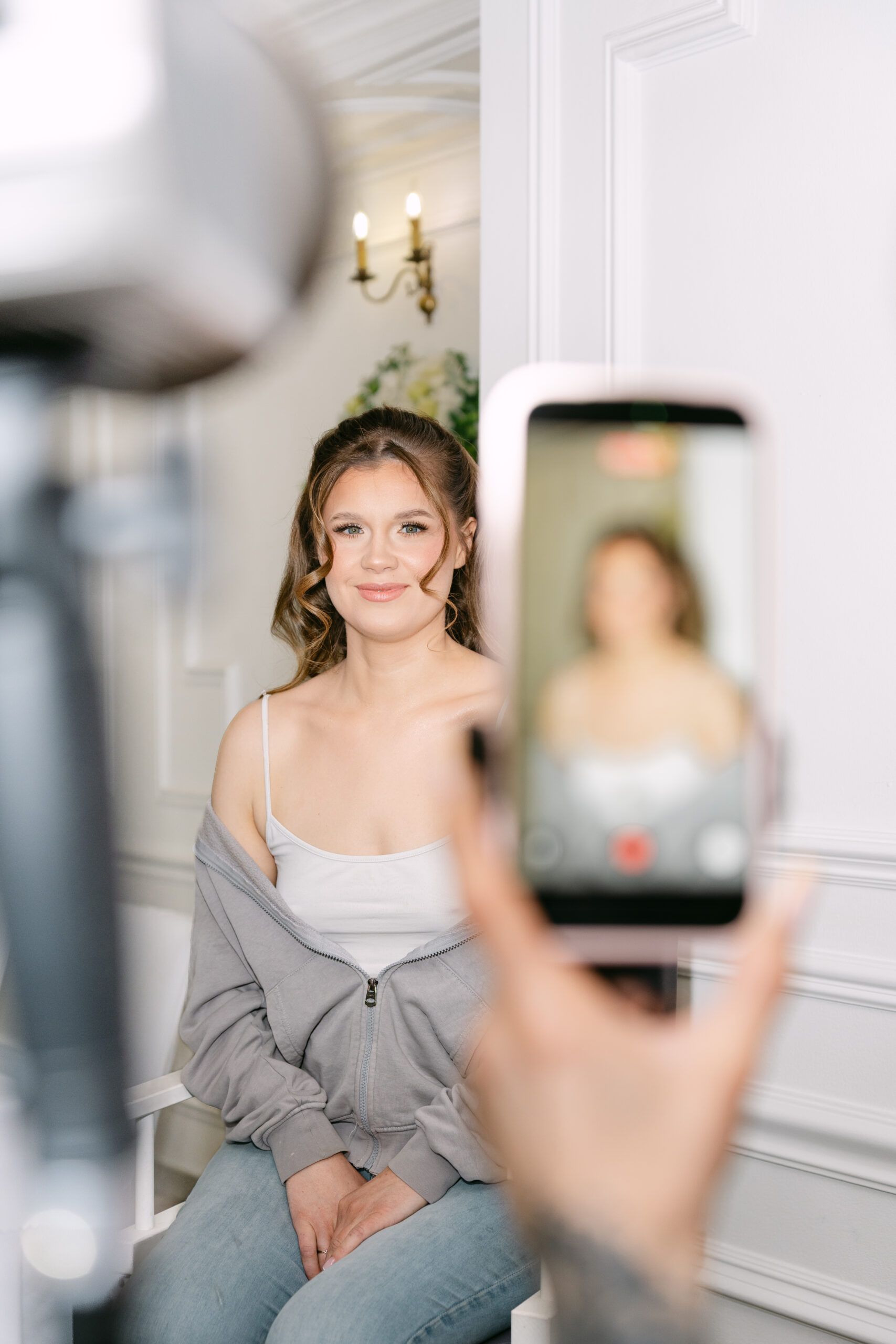 Panna Młoda, Monika, podczas robienia ślubnego makijażu i fryzury. Kadr z przygotowań przedstawiający uśmiechniętą kobietę, nagrywaną smartfonem przez makijażystkę w Dworku Kurowskim.