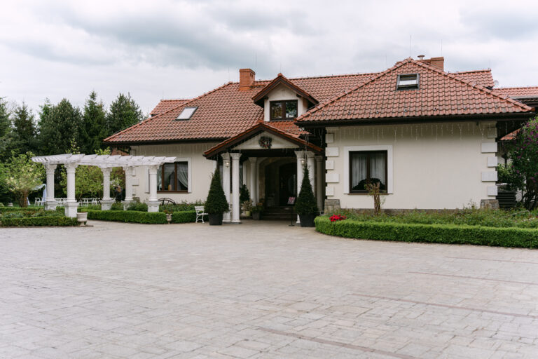 Widok zewnętrzny na Dworek Kurowski – elegancki obiekt na wesele i ślub plenerowy. Szerokie ujęcie budynku z kolumnami, zadbanym podjazdem i zielonym ogrodem w tle.