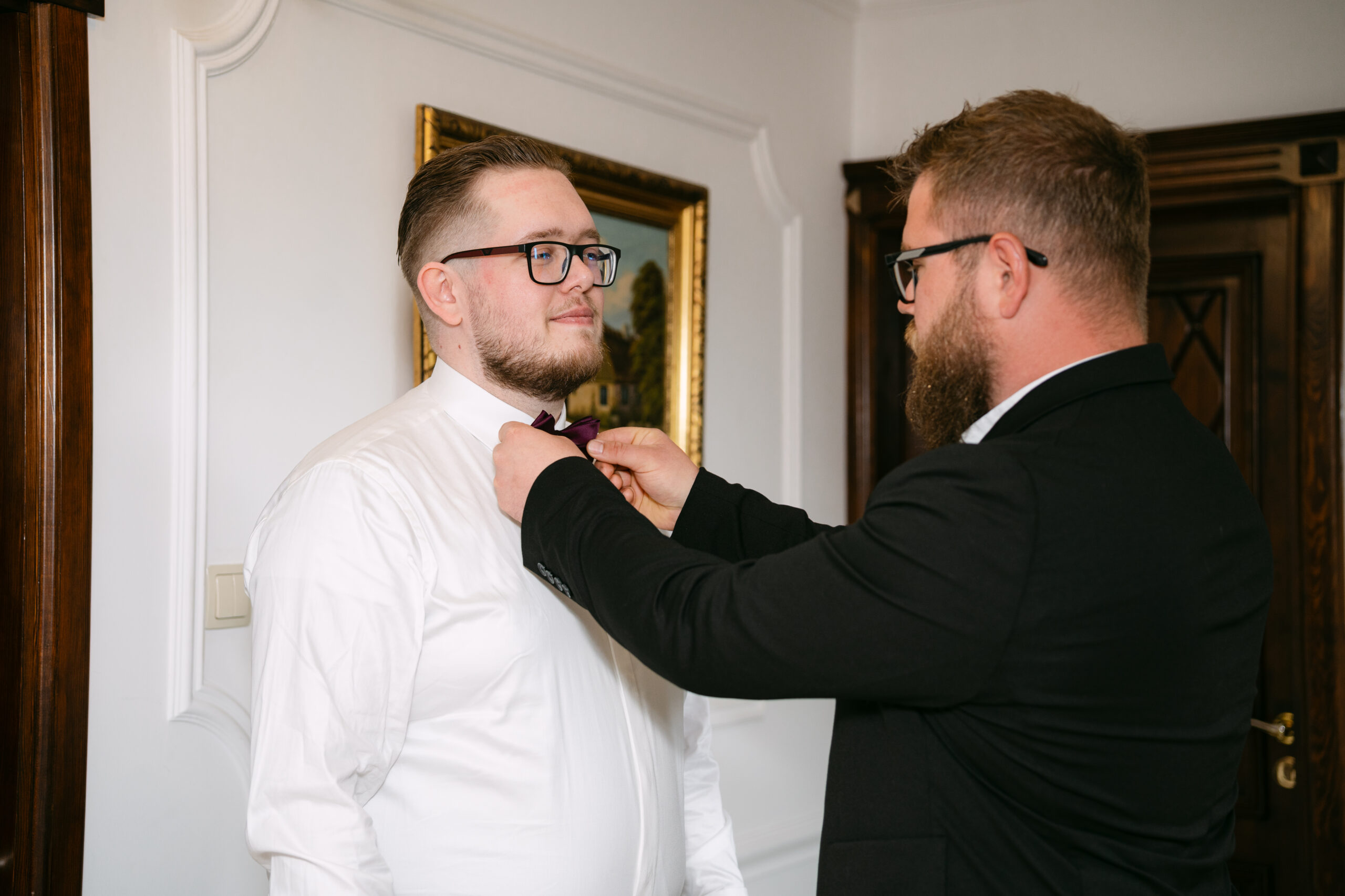 Świadek pomagający Panu Młodemu, Sebastianowi, w poprawianiu bordowej muchy przed ślubem. Bliski kadr z męskich przygotowań ślubnych w Dworku Kurowskim.