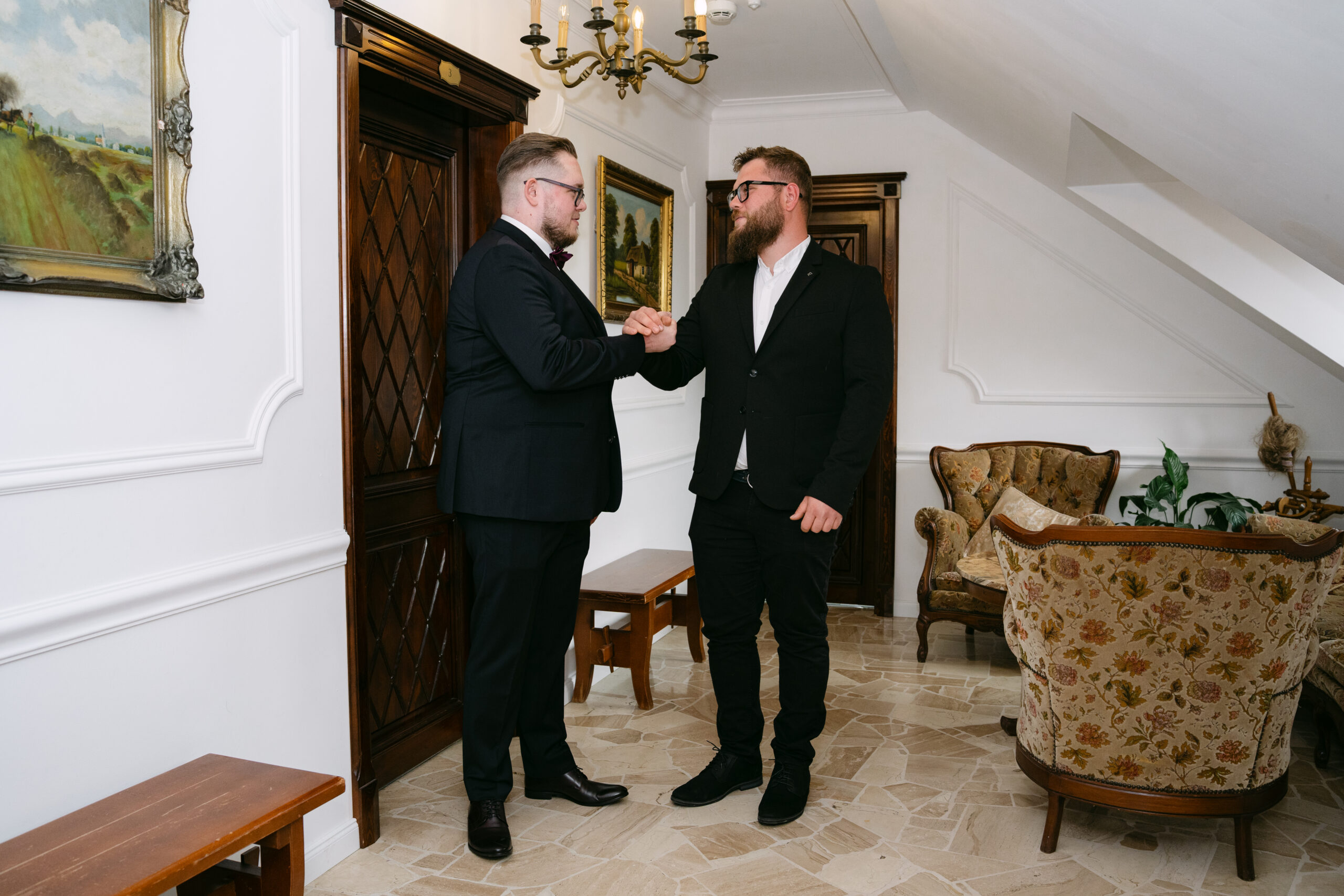 Pan Młody, Sebastian, witający się uściskiem dłoni ze świadkiem w stylowych wnętrzach Dworku Kurowskiego. Męskie przygotowania do ślubu uchwycone w reportażu weselnym.