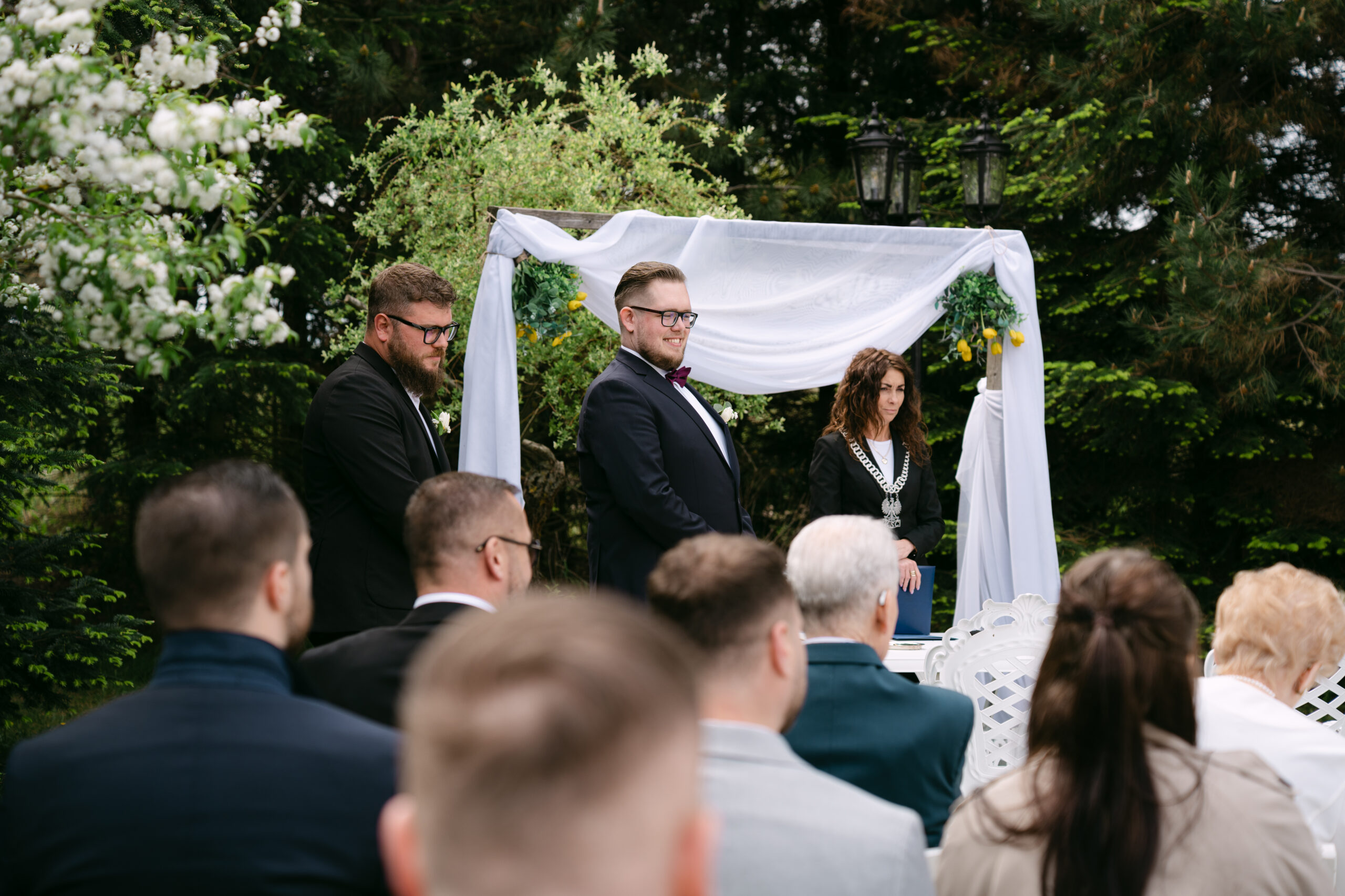 Uśmiechnięty Pan Młody, Sebastian, oczekujący przy łuku ślubnym w ogrodzie na rozpoczęcie ceremonii. Zdjęcie z reportażu ze ślubu plenerowego w Dworku Kurowskim, w tle urzędniczka oraz zgromadzeni goście.