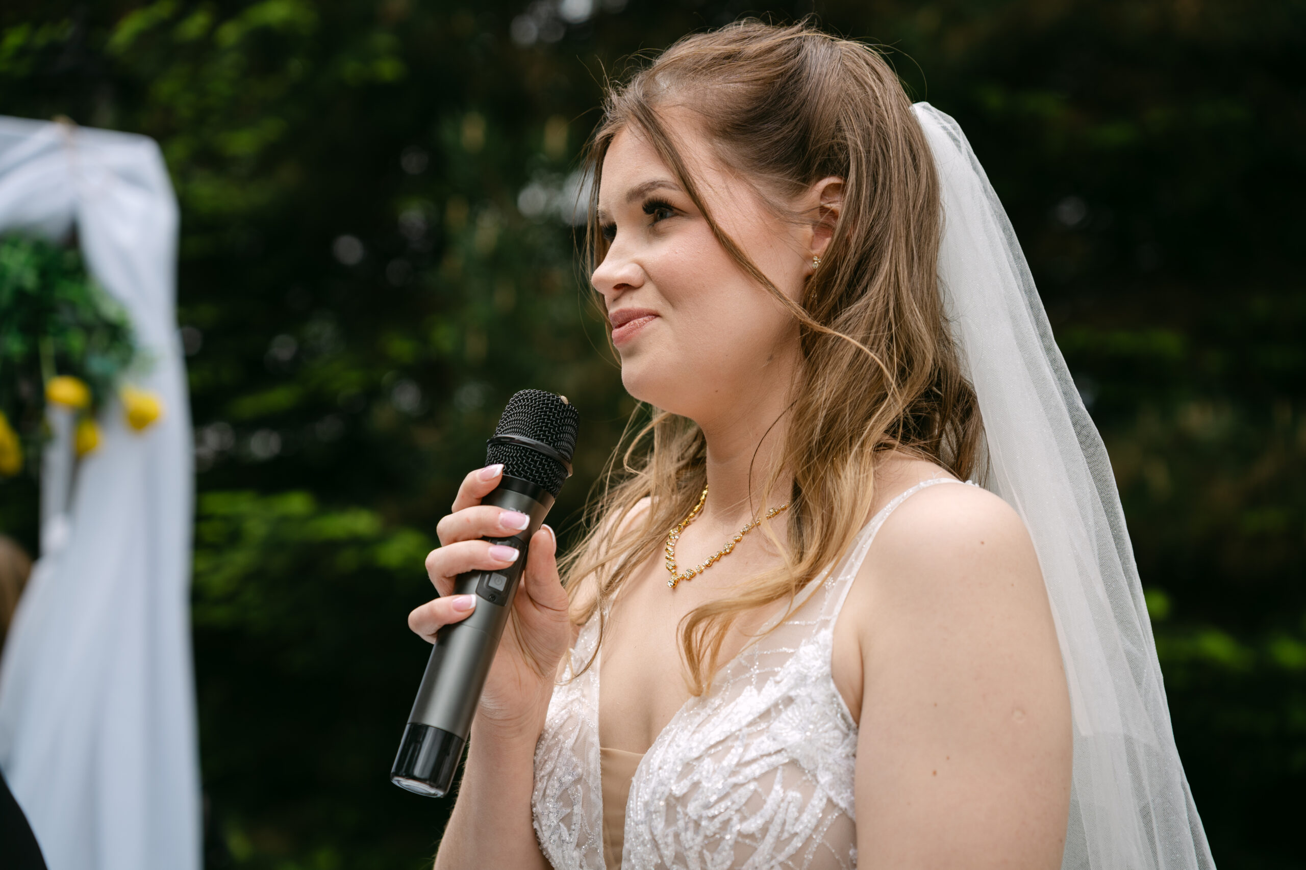 Wzruszona Panna Młoda, Monika, przemawiająca do mikrofonu podczas przysięgi małżeńskiej. Emocjonalny reportaż ze ślubu plenerowego w ogrodzie Dworku Kurowskiego.