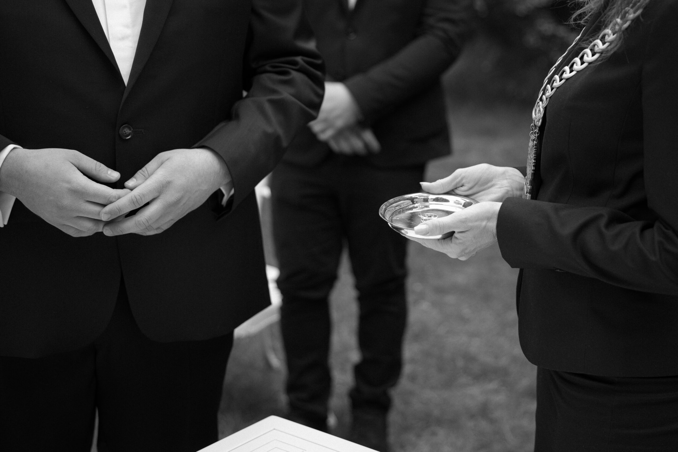 Czarno-biały, bliski kadr na dłonie urzędnika trzymającego obrączki ślubne na tacy podczas ceremonii cywilnej. Emocjonalny reportaż ślubny Moniki i Sebastiana w Dworku Kurowskim.